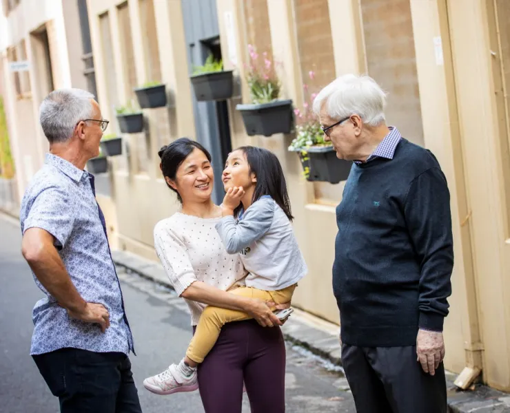 Two older men stand on either side of a young woman holding a small child. They all stand together chatting in a residential alleyway.